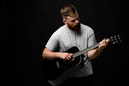Concentrated brutal handsome bearded man in grey t-shirt playing acoustic guitar. Portrait of a male singer and guitarist