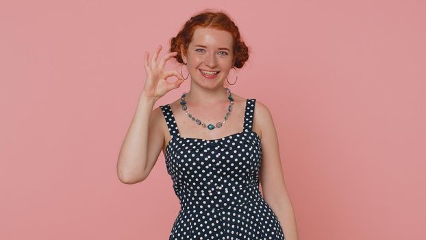 Ok. Happy young woman in polkadot dress looking approvingly at camera showing ok gesture, like sign positive, approve something good. Ginger girl with freckles isolated alone on pink studio background