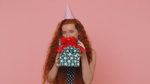 Cheerful young redhead woman opening gift box, unwrapping birthday surprise and expressing great happiness amazement, satisfied with best present bonus. Ginger girl with freckles on pink background