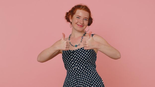 Redhead young woman in polkadot dress raises thumbs up agrees with something or gives positive reply recommends advertisement likes good. Lovely ginger girl isolated alone on pink studio background