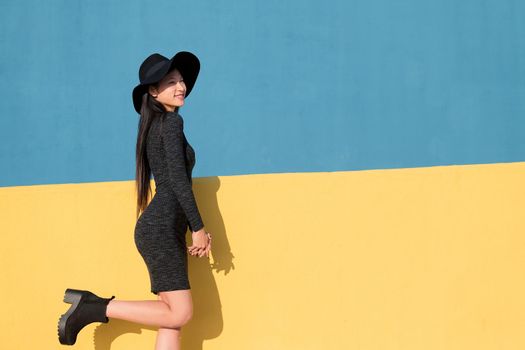 fashionable asian young woman posing happy in a black tight dress and hat on a colorful yellow and blue background, concept of fashion and lifestyle, copyspace for text