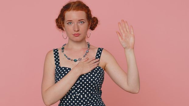 I swear to be honest. Sincere responsible redhead woman raising hand to take oath, promising to be honest and to tell truth keeping hand on chest. Ginger girl with freckles isolated on pink background