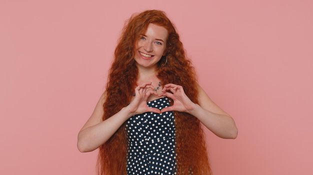 Girl in love. Smiling redhead woman in green dress makes heart gesture demonstrates love sign expresses good feelings and sympathy. Young pretty ginger girl isolated alone on pink studio background