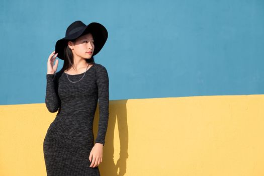 fashionable oriental young woman posing in a black tight dress and hat on a colorful yellow and blue background, concept of fashion and lifestyle, copyspace for text