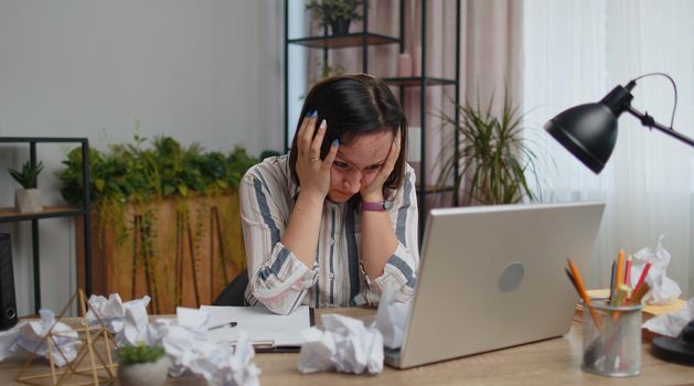 Sad young woman working at home office and brainstorming herself writing down on paper. Dissatisfied businesswoman crumples throwing the written-off sheets of paper with bad ideas and mistakes. Crisis