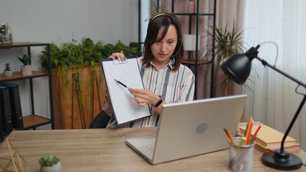 Young business woman sitting at desk at home office wearing headset, freelance worker, call center, support service operator helpline, having talk with client or colleague at laptop computer notebook