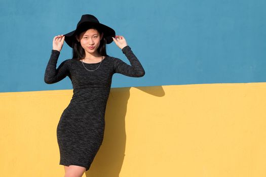 fashionable asian young woman posing in a black tight dress and hat on a colorful yellow and blue background, concept of fashion and lifestyle, copyspace for text
