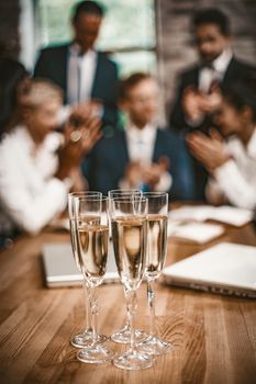 Glasses With Champagne Stand On A Wooden Table In The Foreground, Diverse Business Team Celebrates The Success Of Startup Project And Applauds On Blurred Background, Toned Image