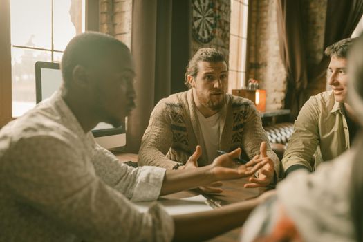 Creative Multi-Ethnic Team Of Freelancers Discussing Startup Project, Colleagues Arguing Each Other Sitting At Table And Gesticulating With Hands In Backlit, Focus On Caucasian Man