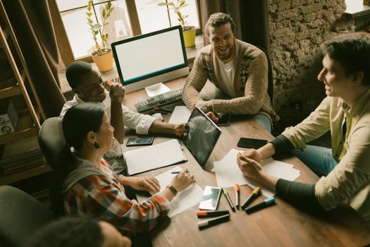 Creative Group Of Employees Works Sitting At Office Desk With White Screen Monitor And Markers On It, Selective Focus On Young Caucasian Man Smiling Near Window, High Angle View