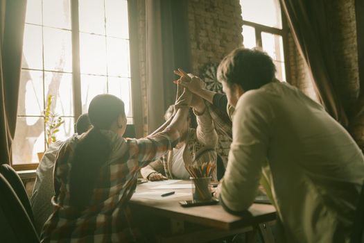 Rear View Of Freelancers Group Bonding Their Hands In High Five Gesture Of Unity, Happy People Join Hands Sittting At Office Table In Backlit