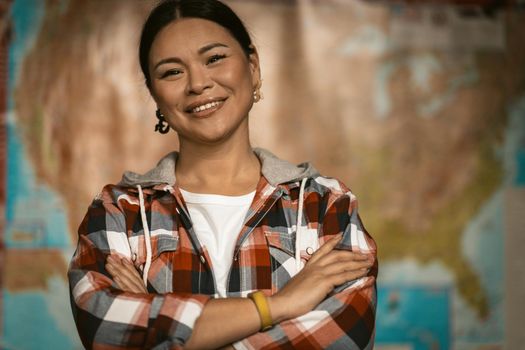 Beautiful Asian Toothy Smiles Standing Against American Map Background, Happy Woman In Casual Clothing Stands With Her Arms Crossed Looking At Camera On Blurred Map Of USA Background