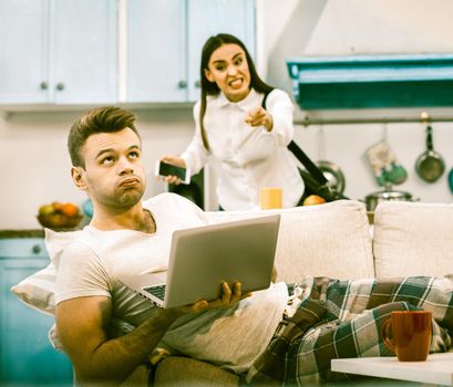 Family Conflict During Home Self-Isolation, Young Wife Is Angry That She Is Bored Of Sitting At Home, Her Husband Makes Displeased Face Lying On Sofa With Computer, Toned Image