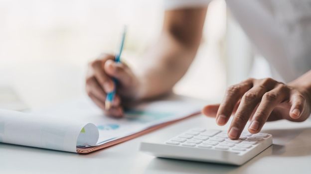 Close up of businessman or accountant hand holding pencil working on calculator to calculate financial data report, accountancy document and laptop computer at office, business concept