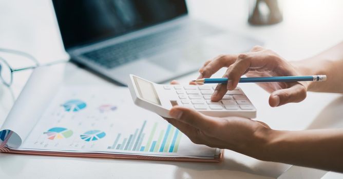 Close up hand of businessman or accountant hand holding pen working on calculator to calculate business data, accountancy document and laptop computer at office, business concept