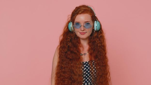 Young redhead woman in dress listening music on headphones, dancing disco fooling around having fun expressive gesticulating hands. Ginger girl with freckles isolated alone on pink studio background