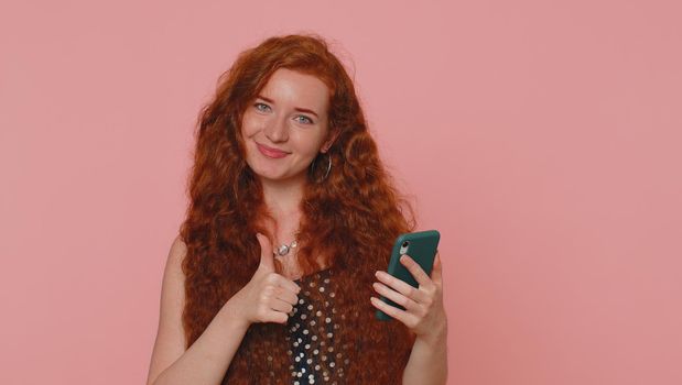 Beautiful red hair woman using mobile phone typing new post on web sms message, browsing, searching addiction of social networks. Young ginger girl studio shot isolated alone on pink wall background