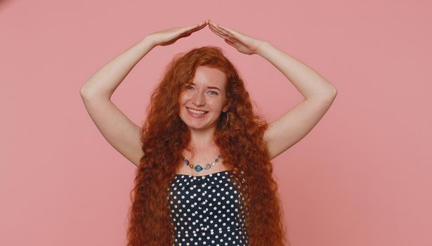 Young redhead woman playing childish catching up game, feeling in safe making roof above head with hands, insurance, security service. Ginger girl with freckles isolated on pink studio background