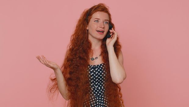 Smiling young redhead woman wearing headset, freelance worker, call center or support service operator helpline, having talk with client or colleague, communication support. Girl on pink background
