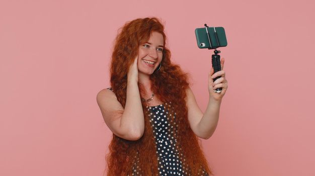 Redhead woman in dress traveler blogger taking selfie on mobile phone selfie stick, communicating video call online with subscribers. Young ginger girl isolated alone on pink studio background