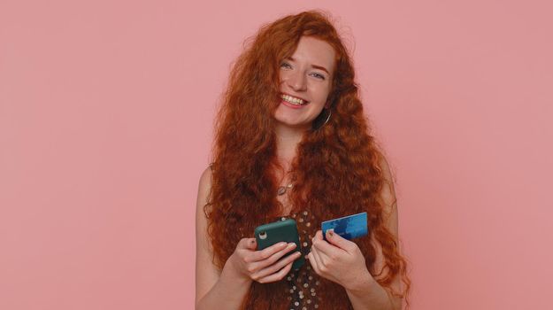 Sincere redhead woman customer using credit bank card and smartphone while transferring money, purchases online shopping, payment. Internet finance. Young girl isolated alone on pink wall background