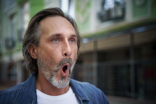 European middle aged man facial muscle exercise outdoors on the street. Mature man dressed up in casual denim jeans shirt stand with open mouth on street. face muscle yoga man outdoors.