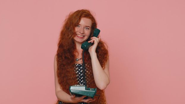 Hey You Call Me Back. Cheerful ginger woman secretary in dress talking on wired vintage telephone of 80s, says hey you call me back. Young redhead freckles face girl isolated on pink studio background