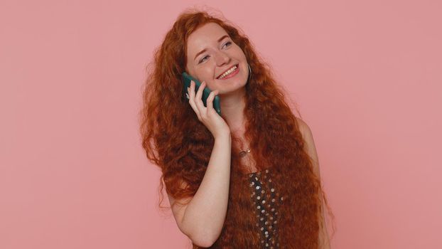 Redhead young woman in polkadot dress having pleasant mobile conversation using smartphone with friends, telling good news. Adult ginger freckles face girl indoors isolated on pink studio background