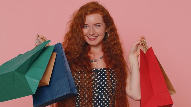 Happy redhead shopaholic woman showing shopping bags, advertising discounts, smiling looking amazed with low prices shopping on Black Friday holidays. Ginger girl indoors isolated on pink background