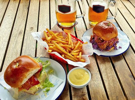 Top view of wooden table with two hamburgers, french fries, mayonnaise and glasses of beer.