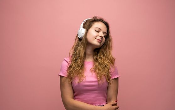 Portrait of charming curly woman in white massive headphones listening to music on isolated pink background. Curly girl in t-shirt smiles