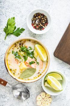 Salmon soup with potatoes, carrot, cream, herbs. Scandinavian/Norwegian fish soup in bowl, cutting wooden board, spoon, rustic white concrete background. Salmon soup for dinner. Top view. Close-up