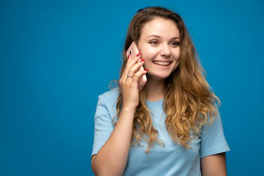 Young smiling happy fun woman in blue basic casual t-shirt talking speak on mobile phone conducting pleasant conversation on blue background studio portrait