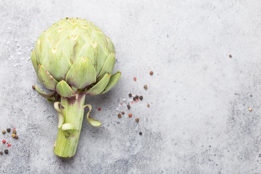 Close-up of raw fresh artichoke with seasonings ready to cook, great as vegetarian food or ingredient for healthy salads and diets, grey stone rustic background, top view. Space for text