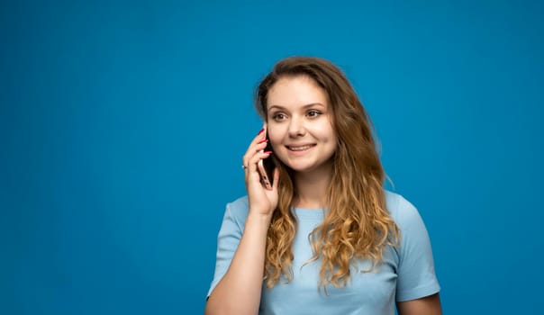 Portrait of a cute happy girl with a curly hair in blue t-shirt talking on mobile phone and laughing isolated over blue background