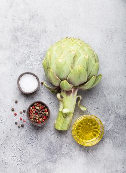 Close-up of raw fresh artichoke with seasonings ready to cook, great as vegetarian food or ingredient for healthy salads and diets, grey stone rustic background, top view