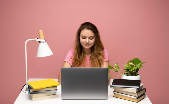 Portrait of a pretty young woman with curly hair studying while sitting at the table with grey laptop computer, notebook. Smiling business woman working with a laptop isolated on a pink background