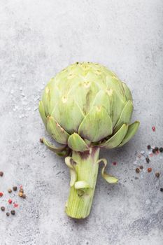 Close-up of raw fresh artichoke with seasonings ready to cook, great as vegetarian food or ingredient for healthy salads and diets, grey stone rustic background, top view