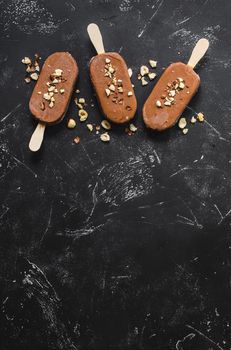 Milk chocolate popsicles with hazelnuts. Ice cream popsicles covered with chocolate, sticks, black stone marble background. Space for text. Top view. Chocolate ice cream bars, nuts. Ice cream dessert