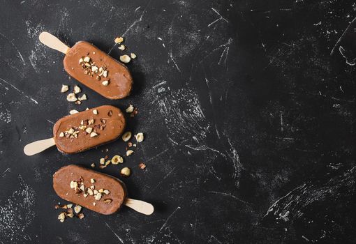 Milk chocolate popsicles with hazelnuts. Ice cream popsicles covered with chocolate, sticks, black stone marble background. Space for text. Top view. Chocolate ice cream bars, nuts. Ice cream dessert