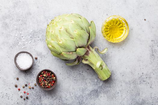 Close-up of raw fresh artichoke with seasonings ready to cook, great as vegetarian food or ingredient for healthy salads and diets, grey stone rustic background, top view