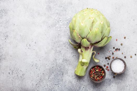 Close-up of raw fresh artichoke with seasonings ready to cook, great as vegetarian food or ingredient for healthy salads and diets, grey stone rustic background, top view. Space for text