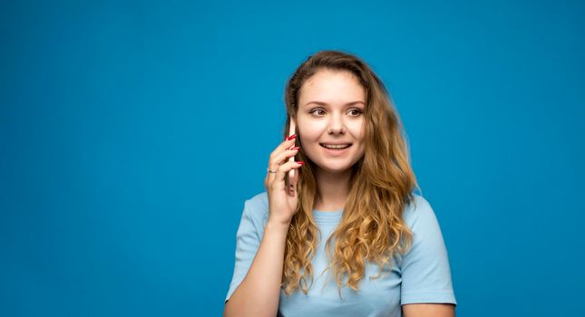 Portrait of a surprised girl with a curly hair dressed in blue t-shirt talking on mobile phone isolated over blue background