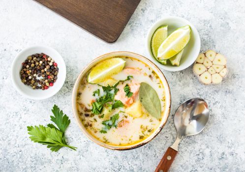 Salmon soup with potatoes, carrot, cream, herbs. Scandinavian/Norwegian fish soup in bowl, cutting wooden board, spoon, rustic white concrete background. Salmon soup for dinner. Top view. Close-up