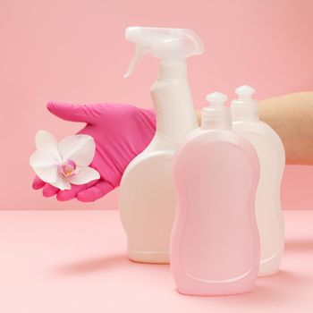 Plastic bottles of dishwashing liquid, glass and tile cleaner, detergent for microwave ovens and stoves and a hand in a nitrile glove with a white orchid flower on a pink background. Washing and cleaning set.