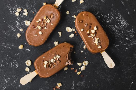 Milk chocolate popsicles with hazelnuts. Close-up. Ice cream popsicles covered with chocolate, sticks, black stone background. Top view. Chocolate ice cream bars, nuts. Dessert. Bitten popsicle