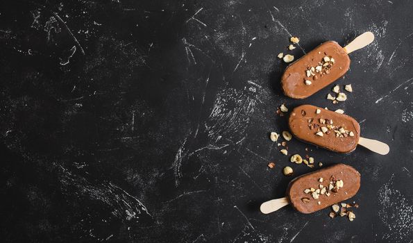 Milk chocolate popsicles with hazelnuts. Ice cream popsicles covered with chocolate, sticks, black stone marble background. Space for text. Top view. Chocolate ice cream bars, nuts. Ice cream dessert