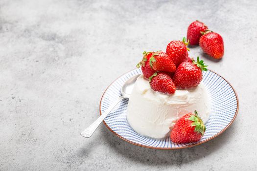 Close-up of fresh Italian cheese ricotta with strawberries on a plate with a spoon, grey rustic stone background, light summer dessert or snack, good for diet or healthy eating with space for text