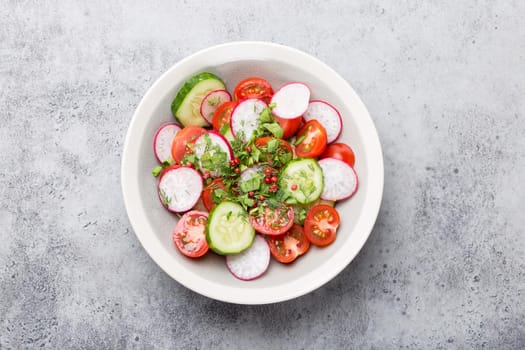 Close-up of fresh summer healthy salad in a bowl made of tomatoes, cucumber, radish and herbs, good for diet, detox or healthy clean eating, grey rustic stone background, top view.