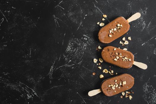 Milk chocolate popsicles with hazelnuts. Ice cream popsicles covered with chocolate, sticks, black stone marble background. Space for text. Top view. Chocolate ice cream bars, nuts. Ice cream dessert